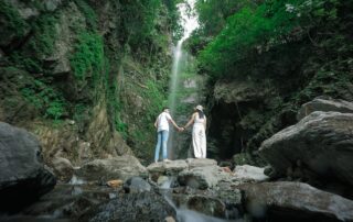 Couple Standing on Rocks on Creek under Waterfall in Forest